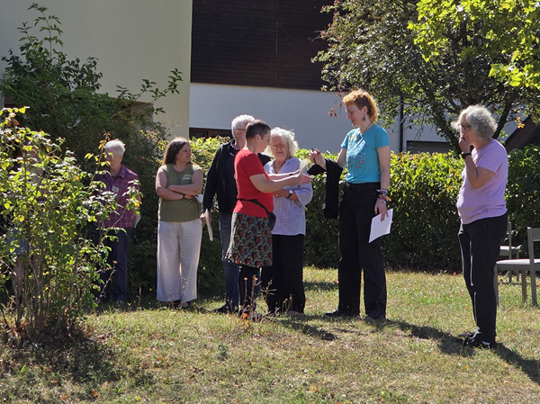 Sieben Menschen im Sonnenschein vor einem Haus. Zwei Personen reden miteinander.