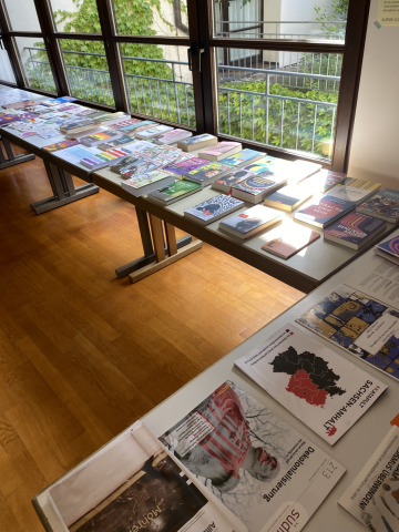 Drei Tische, zu einer langen Tafel zusammengestellt. Darauf sind viele Bücher, Hefte, Sticker und Postkarten zu sehen. 